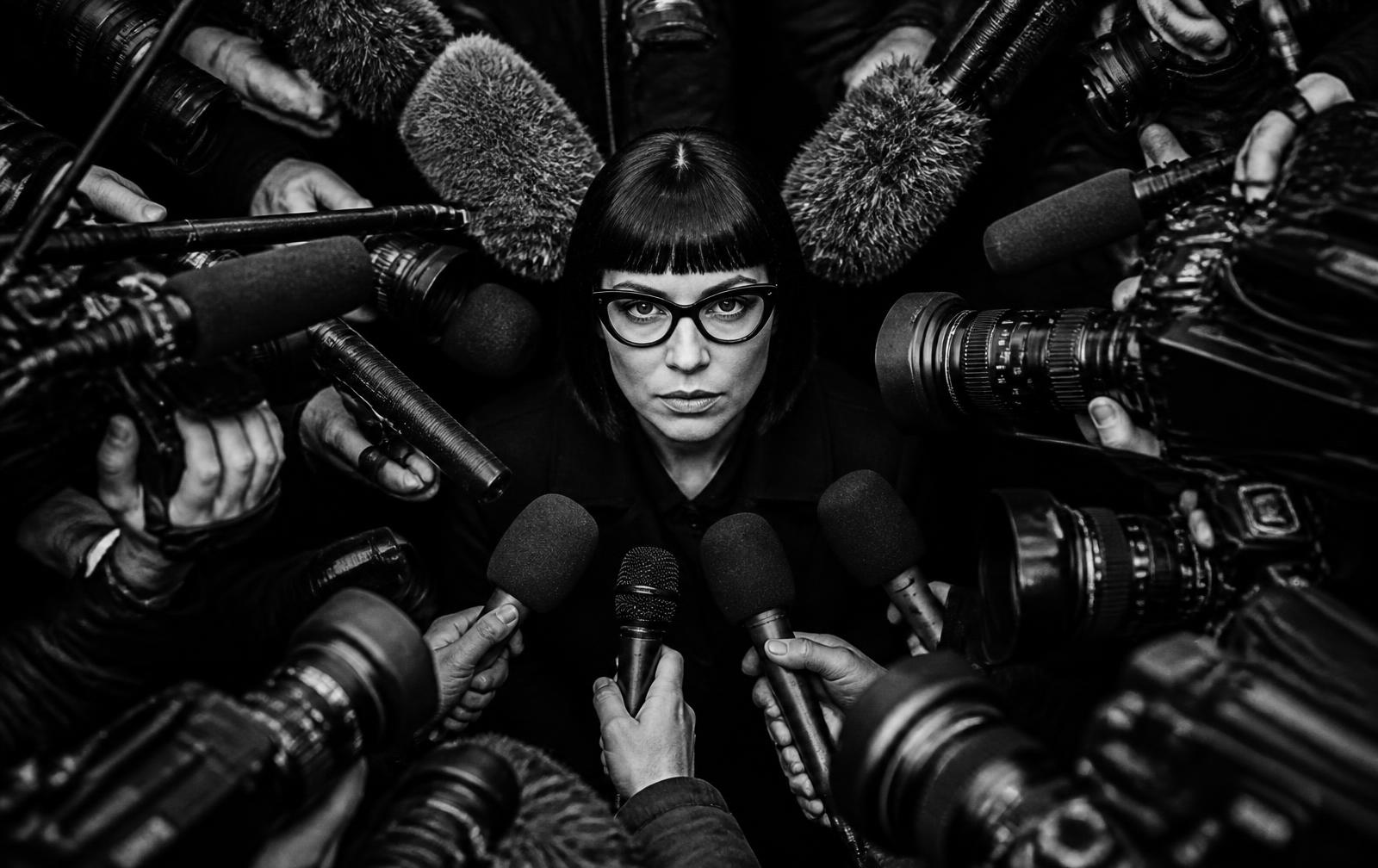 Editorial portrait of a campaign operator in sculptural black tailoring and strong eyewear against a black studio ground, a single red pocket square.