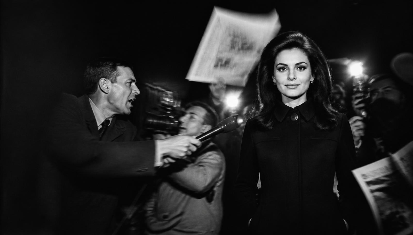 Black and white press scene: one composed media operator walks steadily through the frame carrying a camera while a microphone thrusts in from the right edge, a photographer turns the wrong way and printed pages sweep past in motion blur above a red floor line.